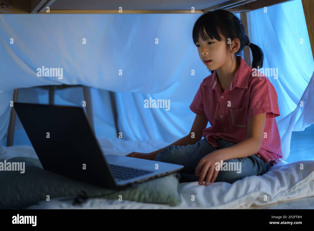 Asian girl child to make a camp to play imaginatively watching a film on laptop in the darkness of the camp in living room at home. Stock Photo