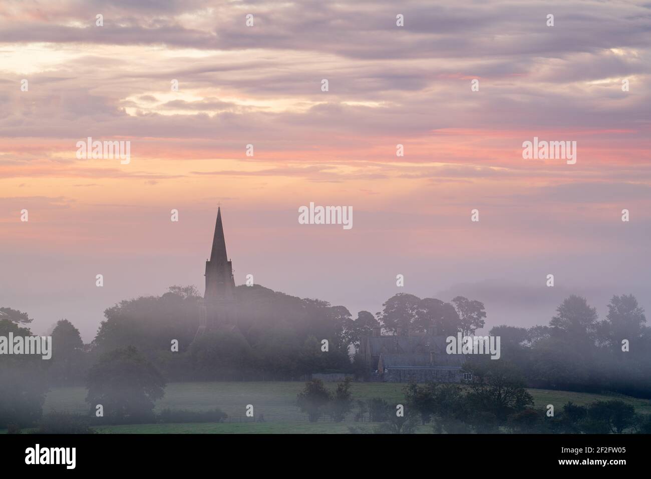 The steeple of St Barnabas Church in Weeton, Lower Wharfedale, is visible above a blanket of summer mist during a pastel August sunrise. Stock Photo