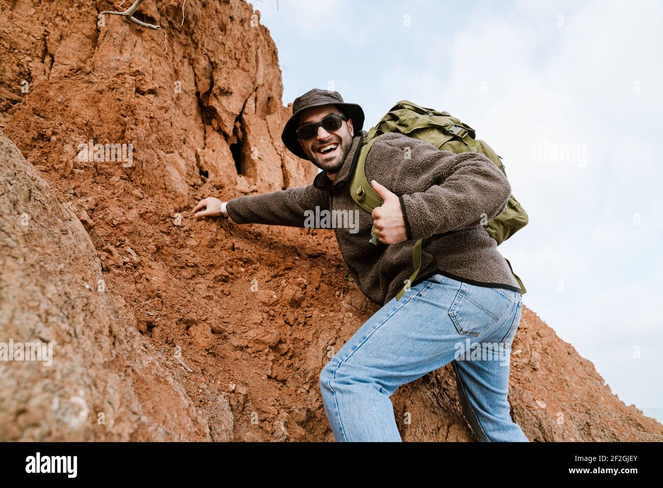 Happy unshaven man showing thumb up while climbing on hill with backpack outdoors Stock Photo