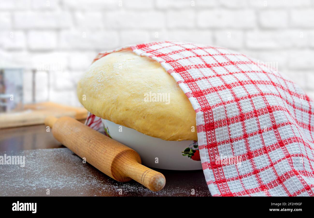 Raw yeast dough covered with a towel in a bowl on the floured kitchen table, recipe idea. Concept home baking or making dough. Stock Photo