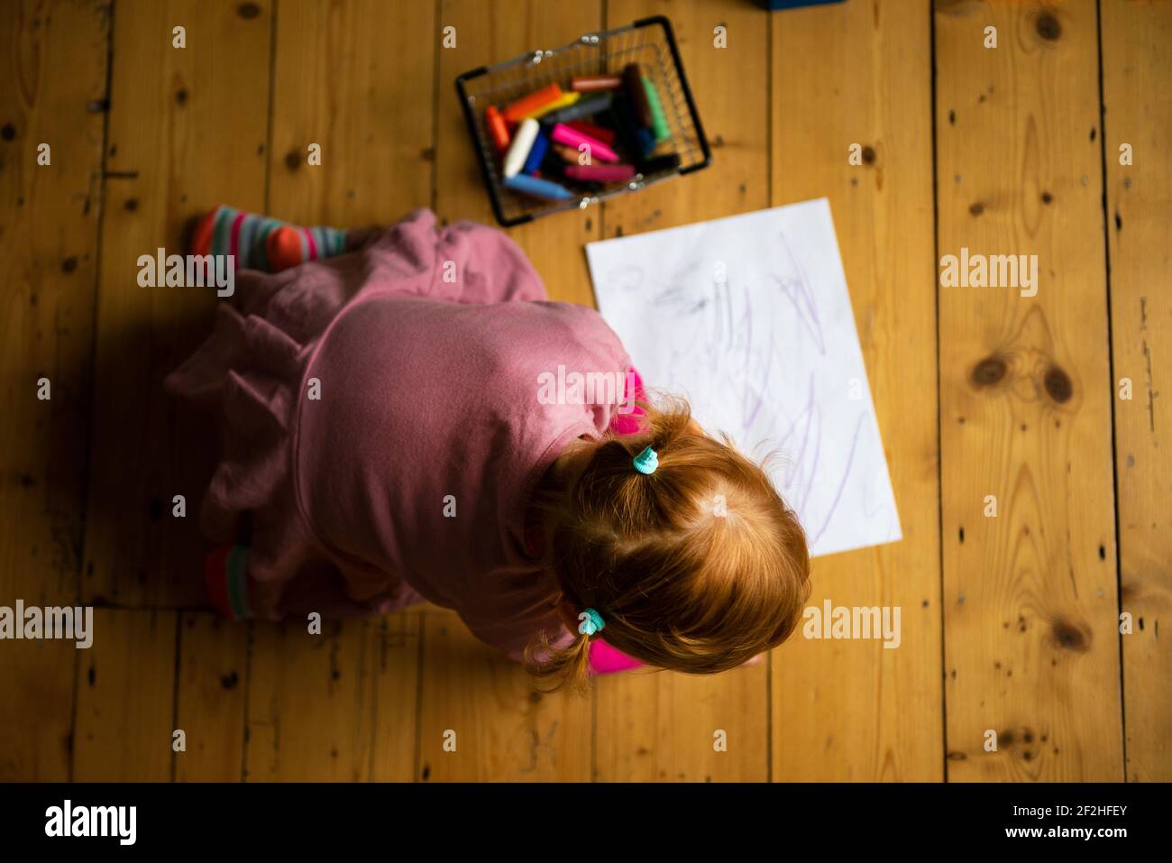 A little girl dressed in pink and with her hair up in pigtails sits on a wooden floor drawing on white paper with colourful crayons. Stock Photo