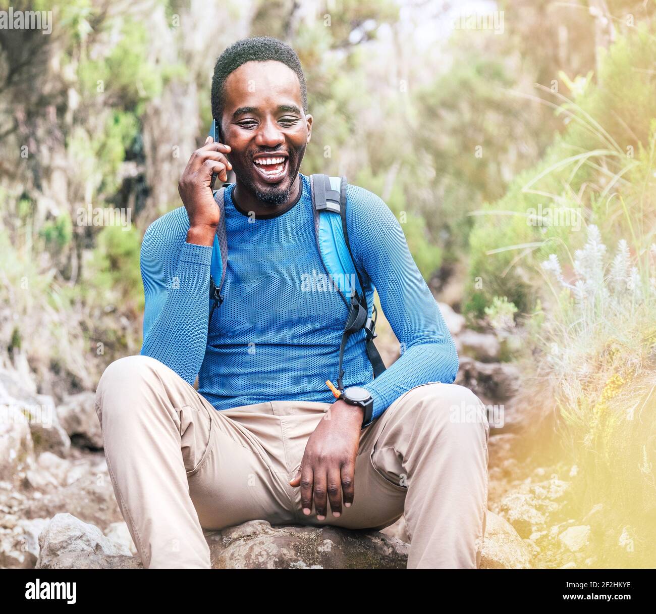 Young African ethnicity man talking via cellphone with somebody and cheerfully laughing as he having a hiking walk in the tropical forest. Happy peopl Stock Photo