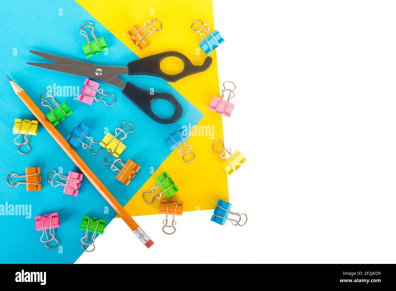 Stationary flat lay on blue and yellow paper with white background and empty space on one side. Lots of small colored paper clips, little scissors, an Stock Photo
