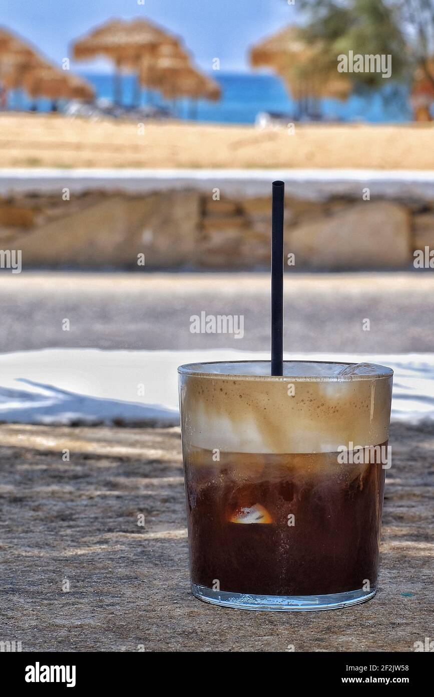 Closeup of an iced coffee with a straw in front of the mylopotas beach in Ios Greece Stock Photo