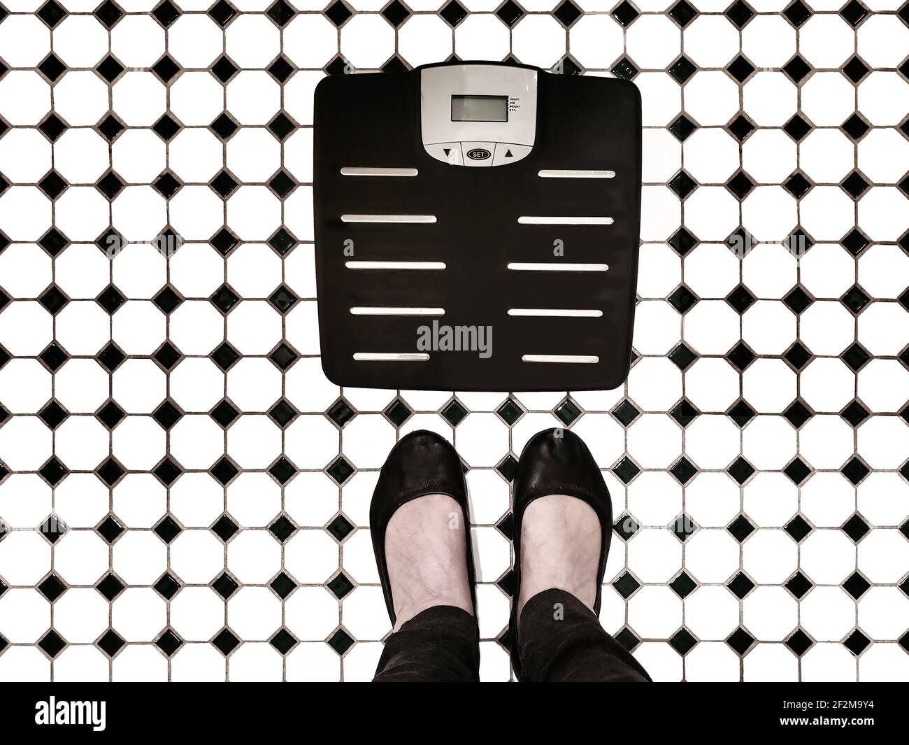 Body weight scales on tiled bathroom floor with woman's feet beside it like shes going to step on them - top view Stock Photo