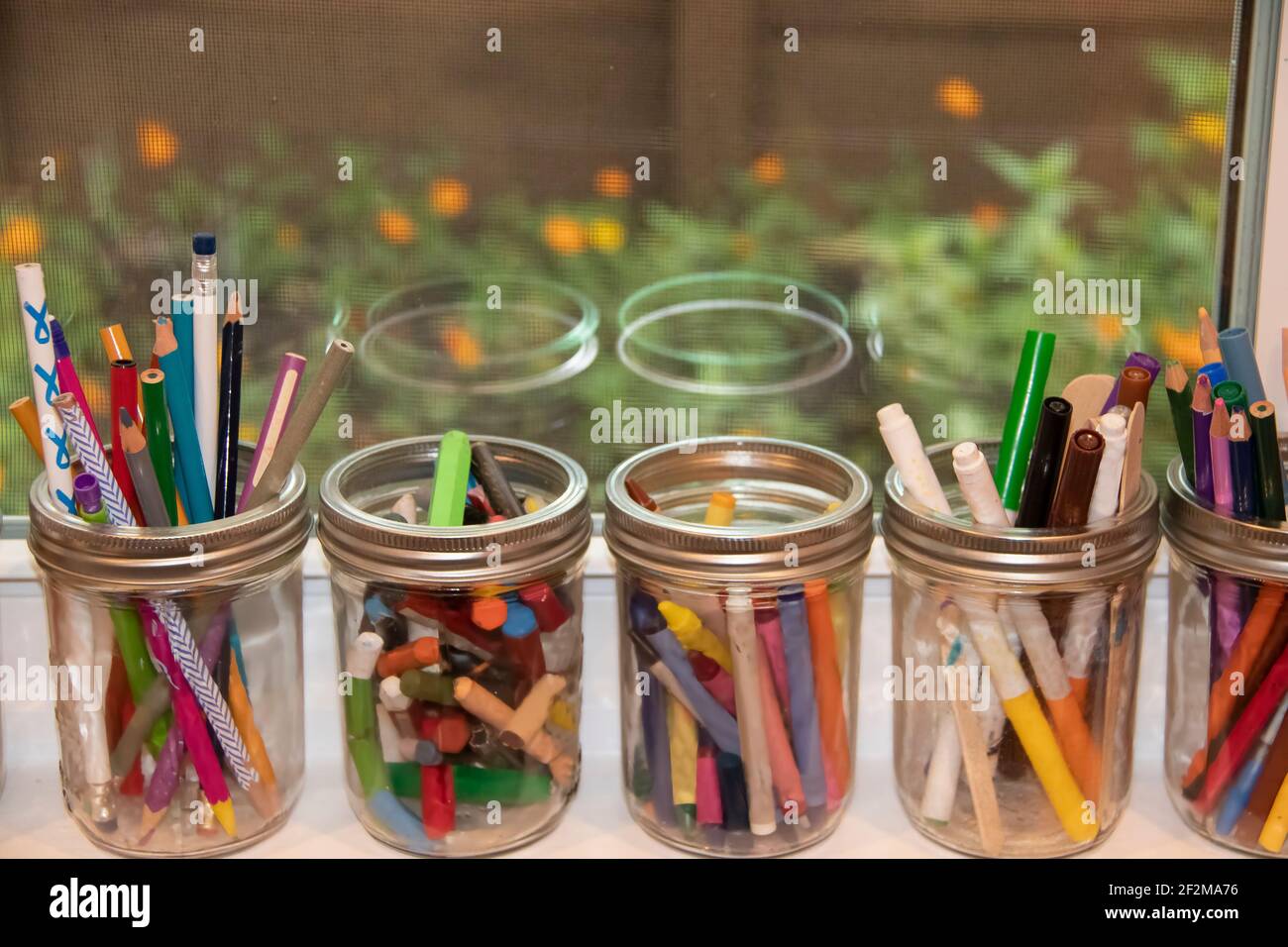 Canning jars filled with crayons, pastels and markers sitting on an open windowsill with flowers outside seen through a window screen Stock Photo