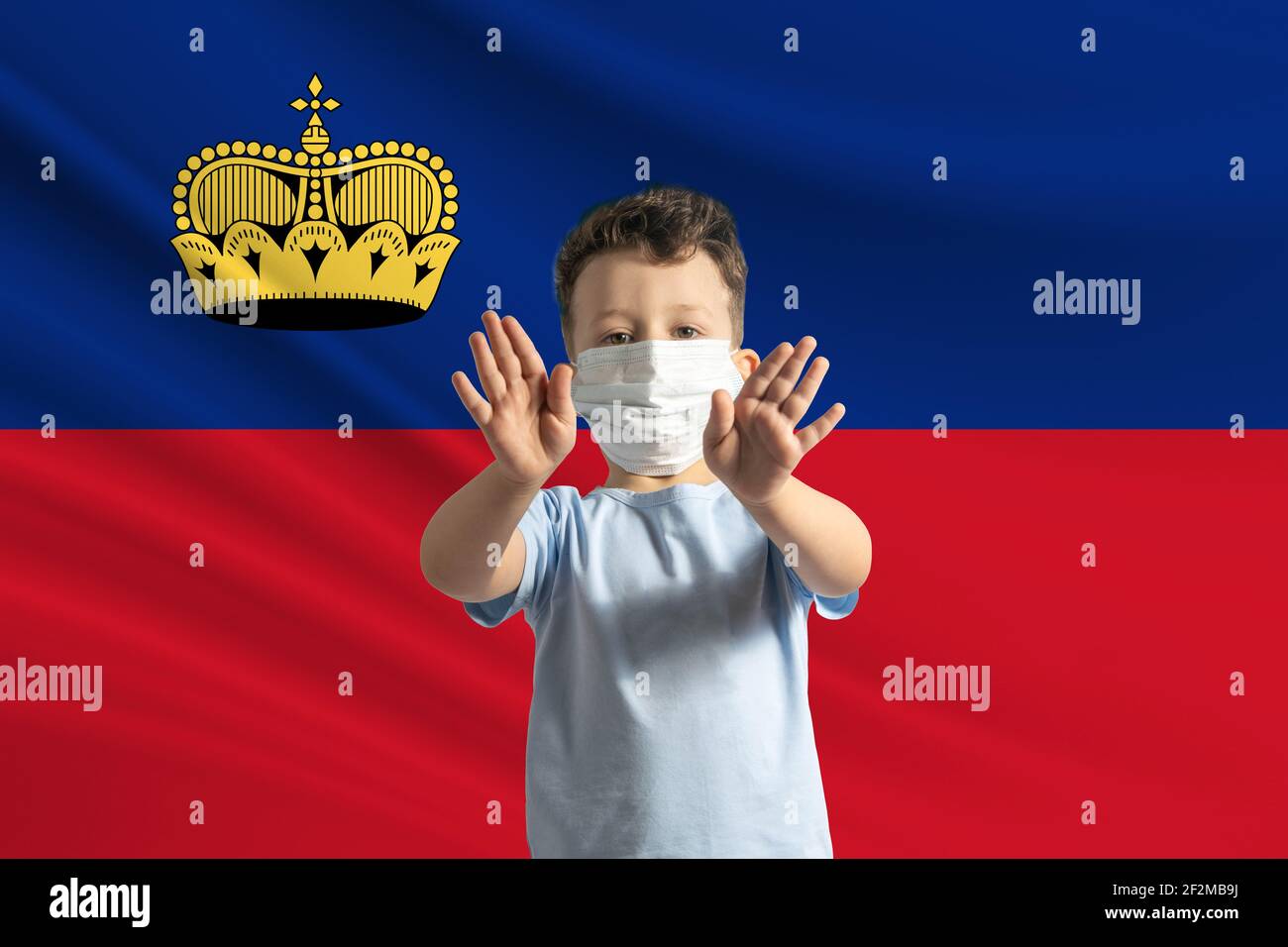 Little white boy in a protective mask on the background of the flag of Liechtenstein. Makes a stop sign with his hands, stay at home Liechtenstein. Stock Photo