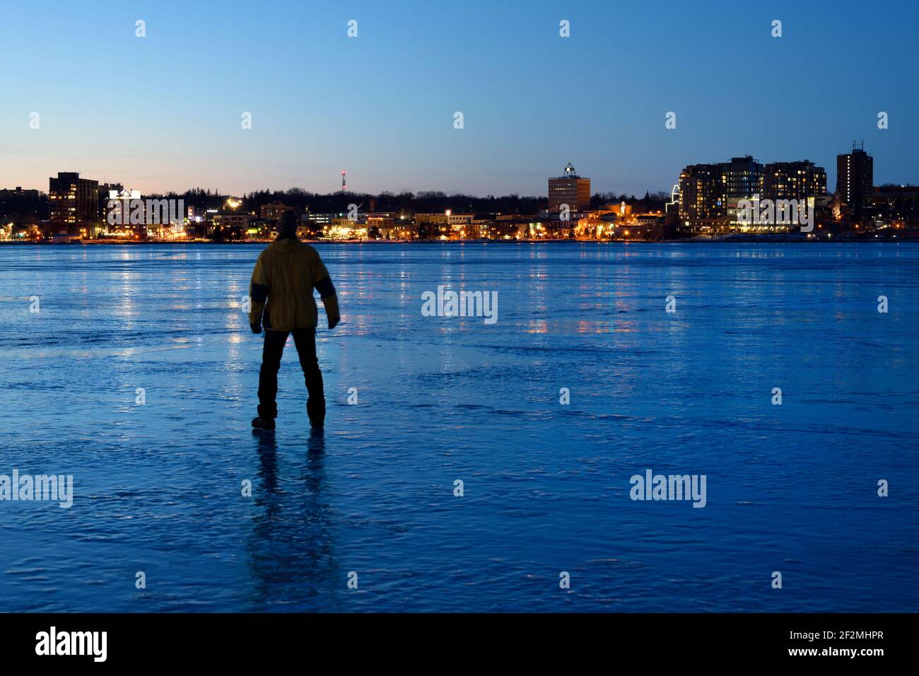 Man watching city light reflections of Barrie Canada on ice of frozen Kempenfelt Bay at blue hour twilight in winter Stock Photo