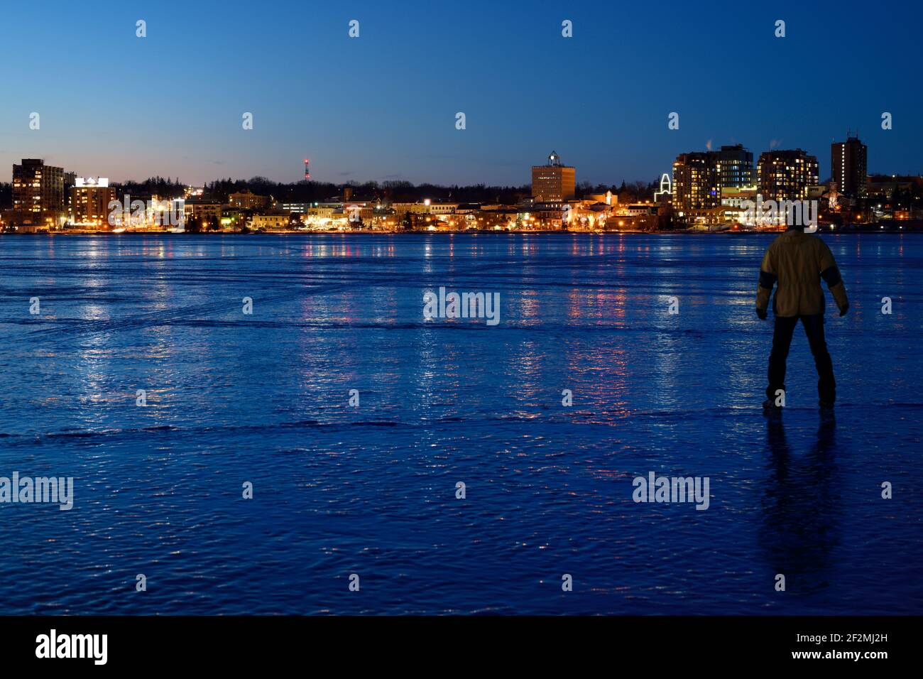 Man standing on thick ice of frozen Kempenfelt Bay at blue hour twilight in winter with lights of downtown Barrie Ontario Canada Stock Photo