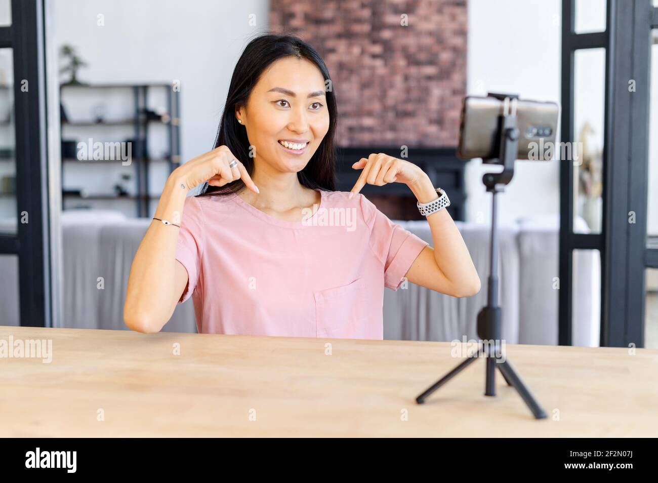 Asian female blogger smiling, sitting at the desk, at home, talking in front of mobile phone standing on a tripod, giving her expert opinion, recommending, shows with her hands to an copy space Stock Photo