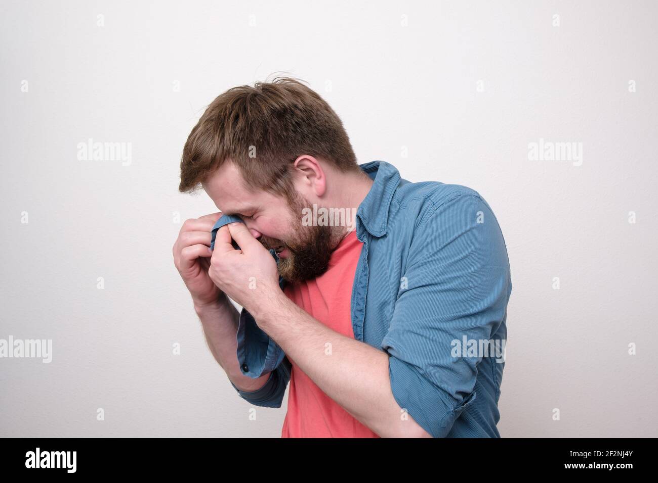 Caucasian bearded man is very upset, he sobbing and wipes away tears with his shirt. White background. Stock Photo