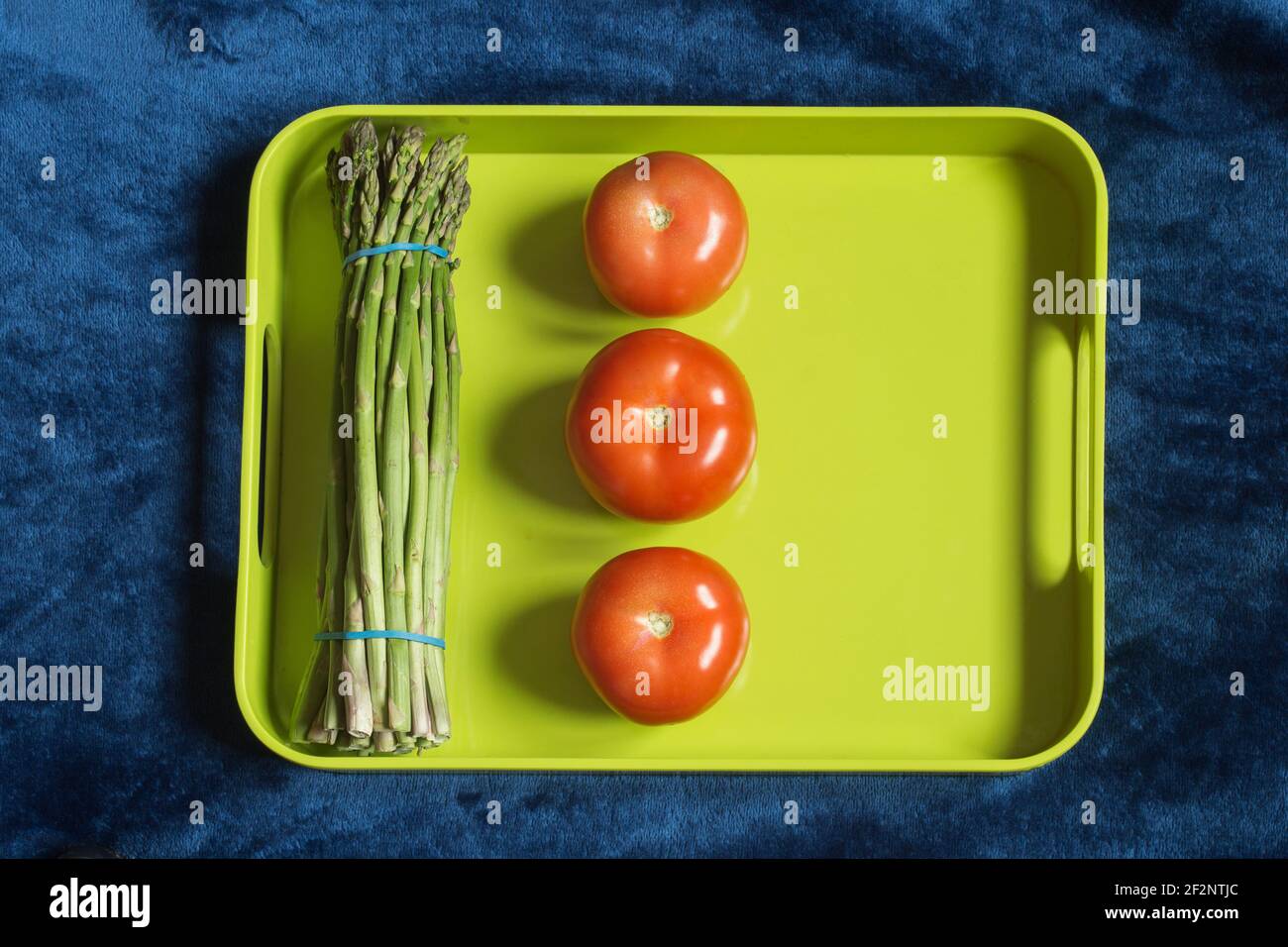 A green food tray with alienated tomatoes and a bunch of wild asparagus on a blue fabric texture background. Food, fruit, and vegetables. Stock Photo