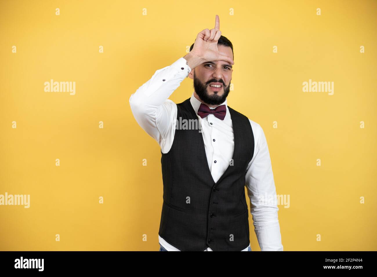 Young man with beard wearing bow tie and vest making fun of people with fingers on forehead doing loser gesture mocking and insulting. Stock Photo