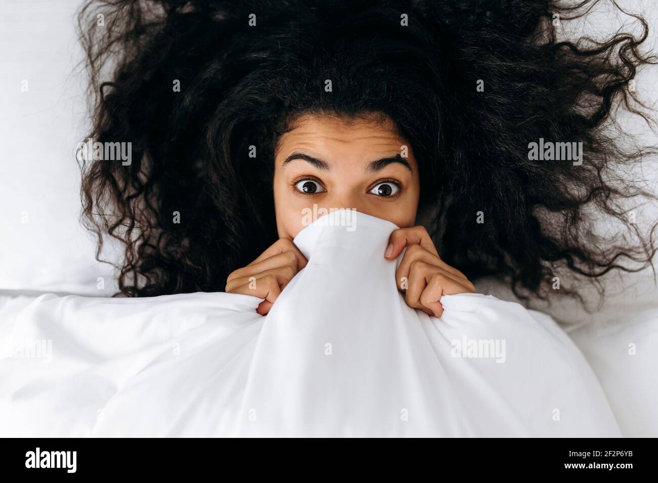 Top view on funny african american girl with curly hair, under white  blanket. Multiracial girl looking