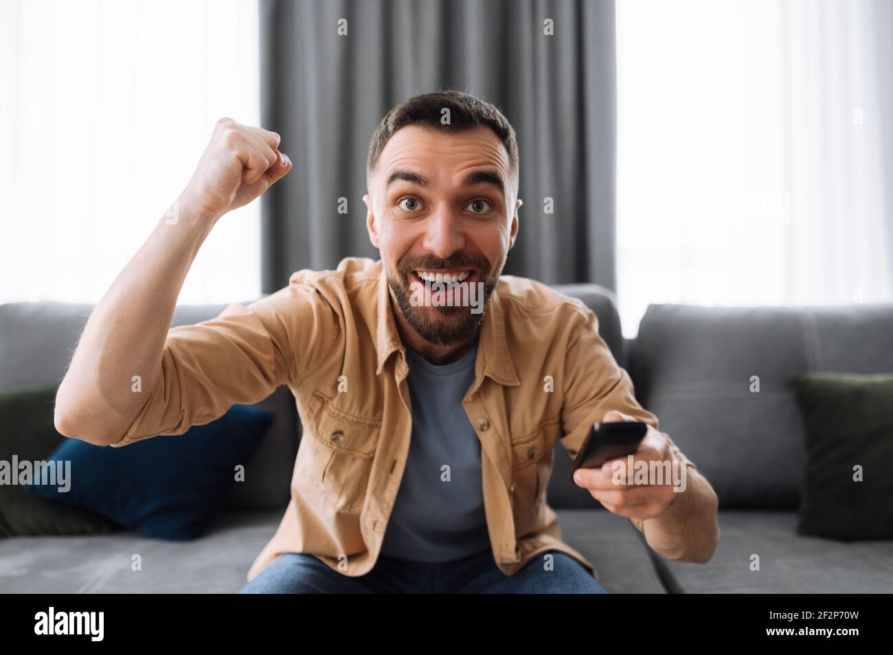 Excited joyful man sitting on the sofa, watching on tv sport channel. Cheerful emotional guy support the team, screaming, rejoices of successful game, football fan Stock Photo