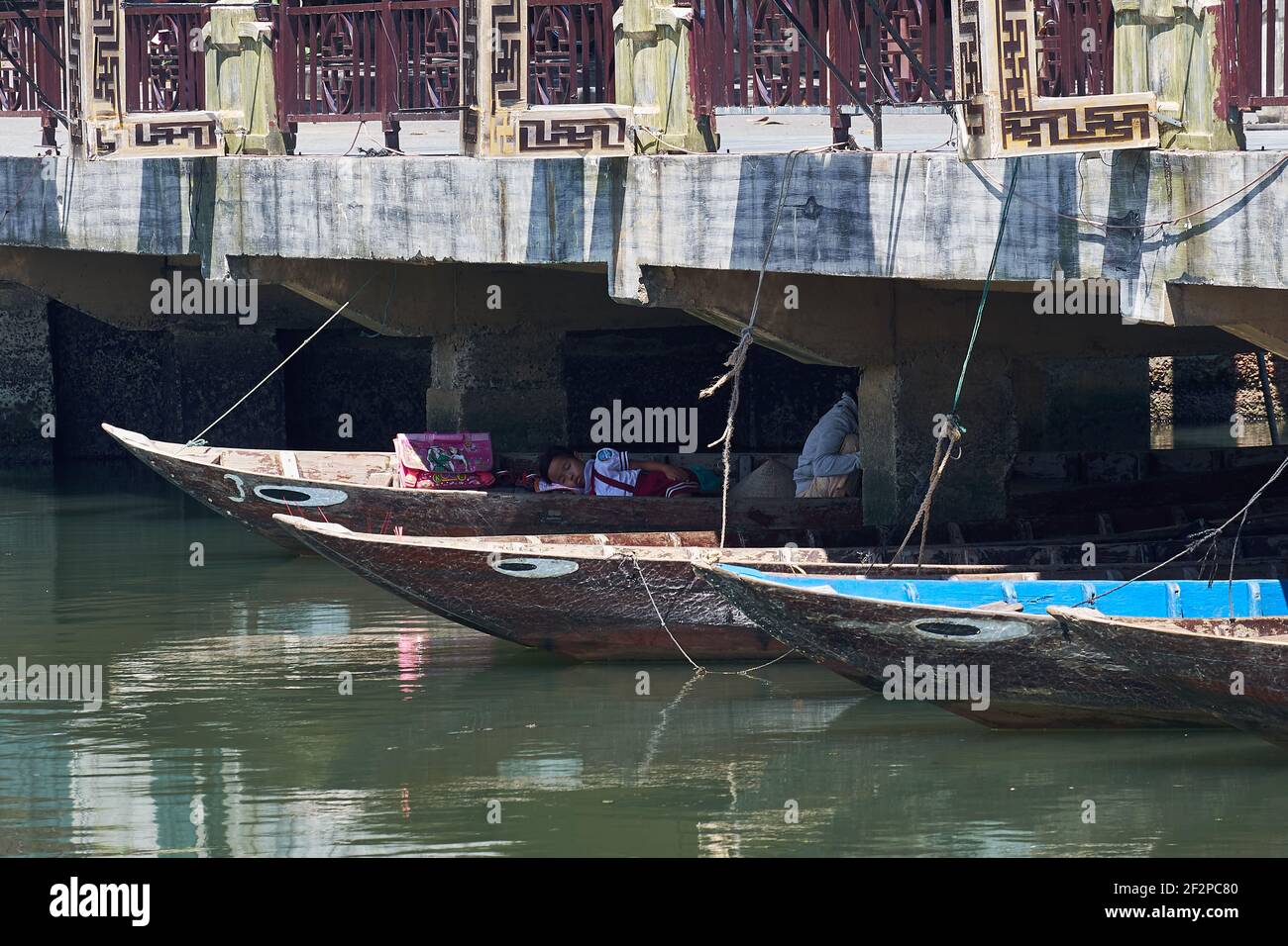 Young girl in school clothes having an afternoon nap in her mother’s boat in the shade underneath the Bridge of Lights (Cau An Hoi) in Hoi An, Vietnam Stock Photo