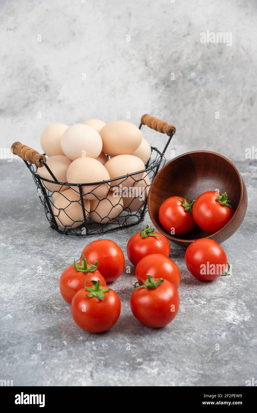 Wicker basket of raw organic eggs and bowl of tomatoes on marble background Stock Photo