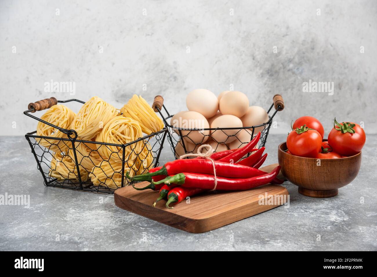Basket of raw noodles, tomatoes, chili peppers and eggs on marble surface Stock Photo