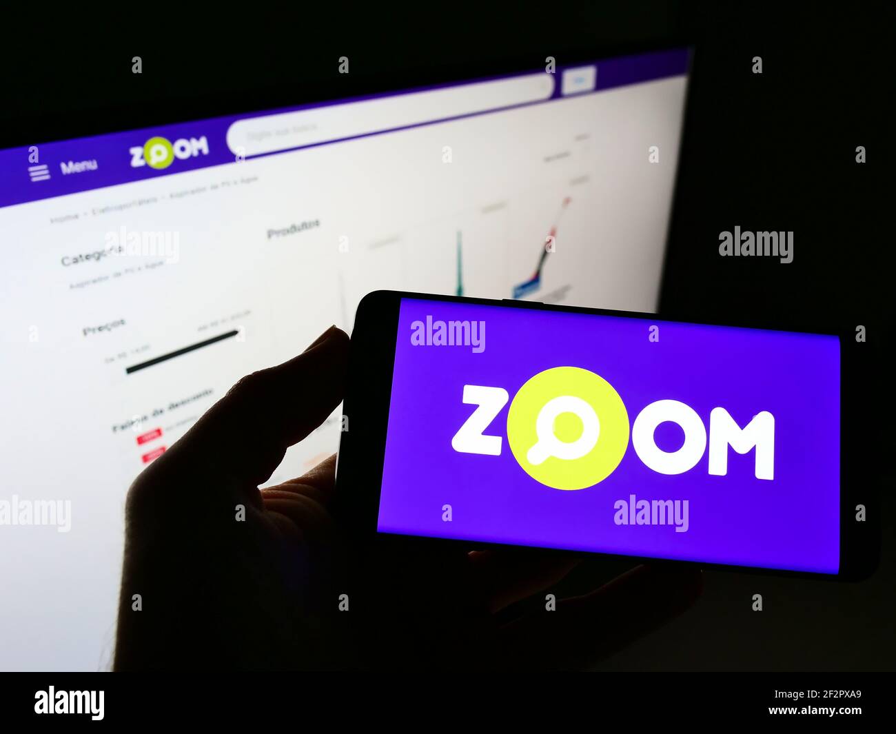 Person holding cellphone with business logo of Brazilian online shop Zoom Brasil on screen in front of web page. Focus on phone display. Stock Photo
