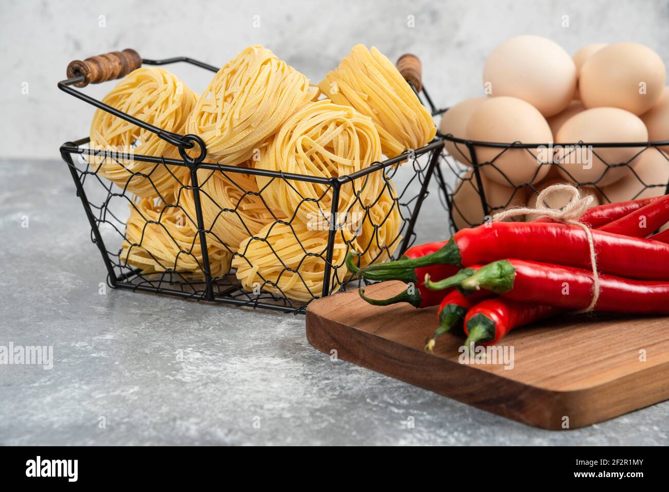 Basket of raw noodles, chili peppers and eggs on marble surface Stock Photo