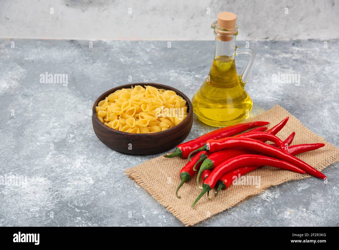 Wooden bowl of raw pasta with red chili peppers and oil on marble background Stock Photo