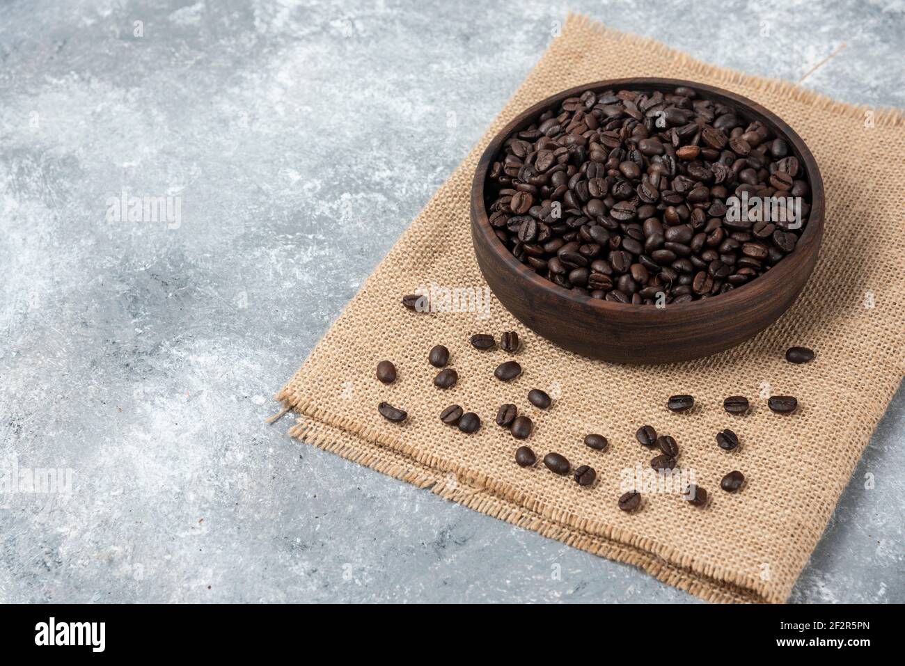 Wooden bowl of dark roasted coffee beans and burlap on marble surface Stock Photo