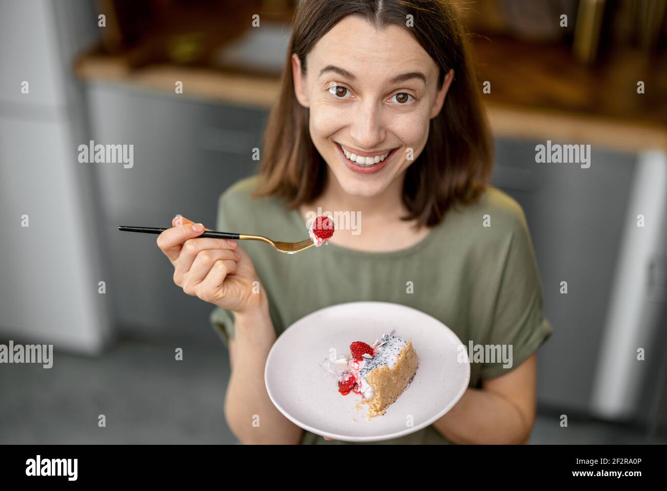 Young woman enjoys a sweet dessert, having a snack on a kitchen at home. Home made recipe of cheesecake with raspberry. Healthy cake. Looking and smiling at camera. Top view. Stock Photo