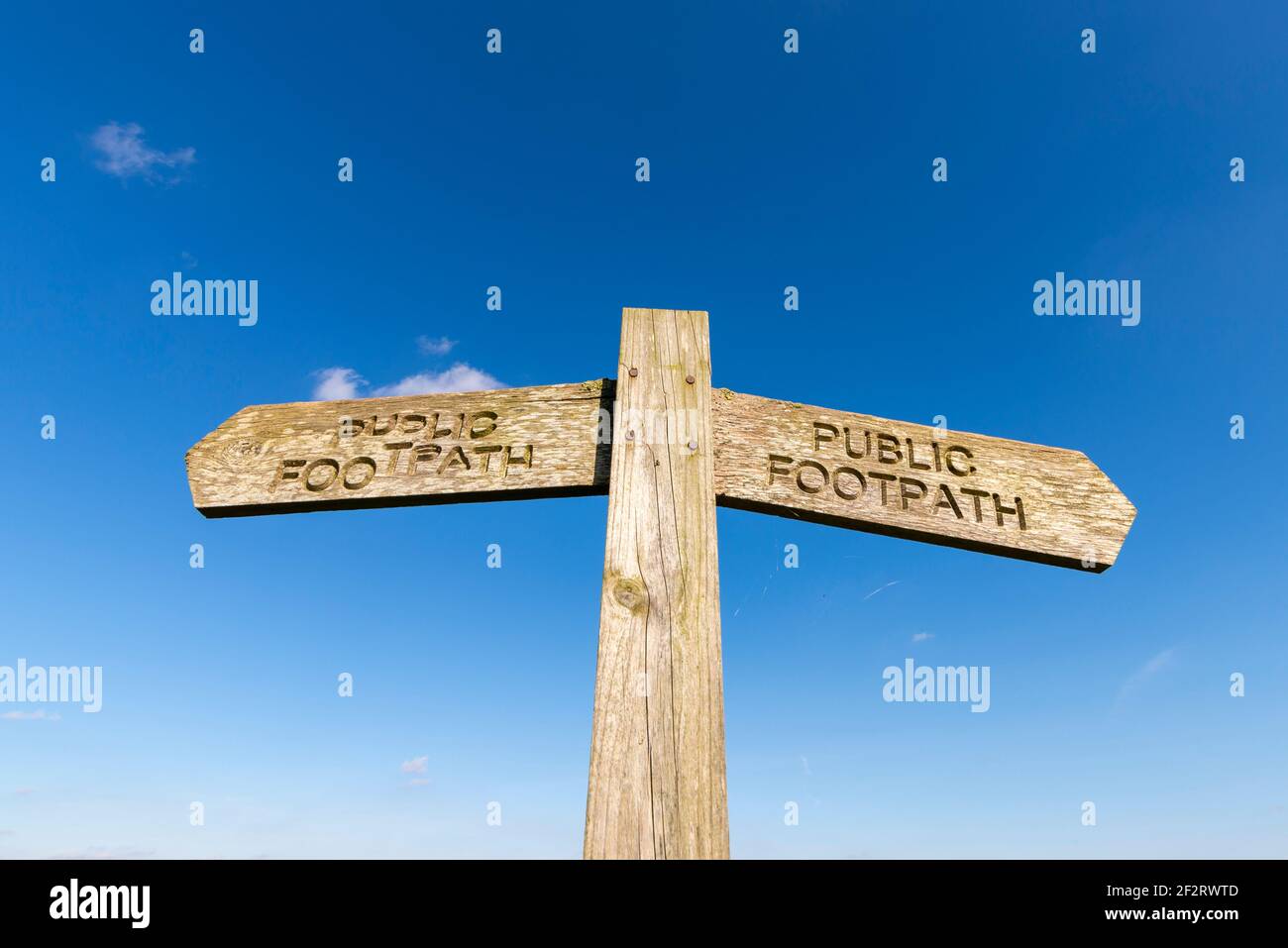 Old wooden public footpath sign in a rural area near Stambridge in the Essex countryside. Isolated against blue sky. Text not straight Stock Photo