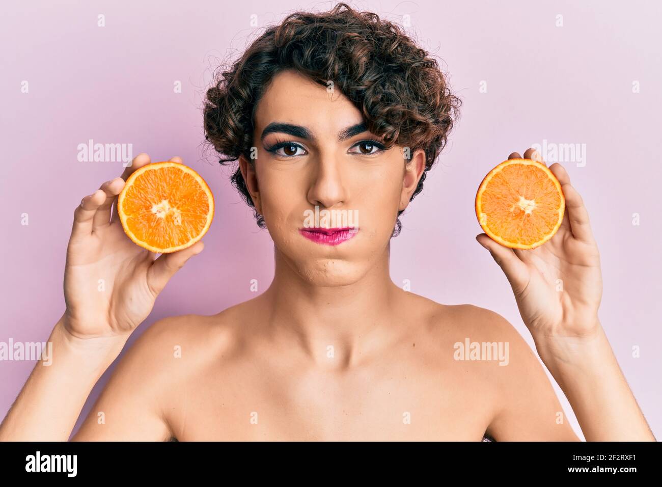 Young man wearing woman make up holding fresh orange puffing cheeks with funny face. mouth inflated with air, catching air. Stock Photo