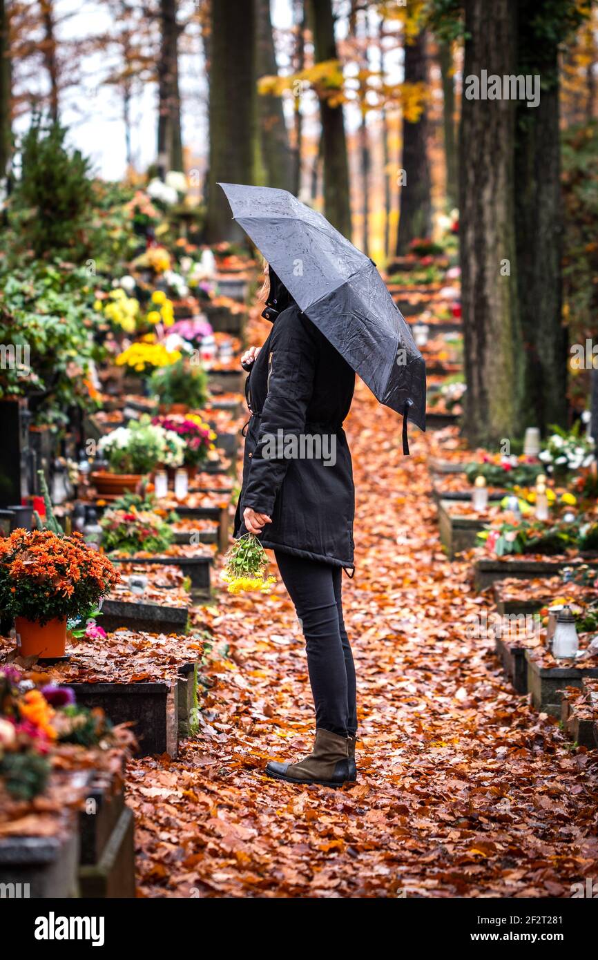 Sad woman with umbrella is mourning for dead person at grave in cemetery. Lonely mourner in autumn graveyard in rain Stock Photo