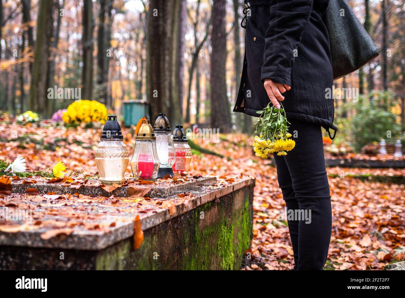 Mourning woman holding flowers in hands and standing at grave in cemetery. Woman wearing black clothing at graveyard Stock Photo