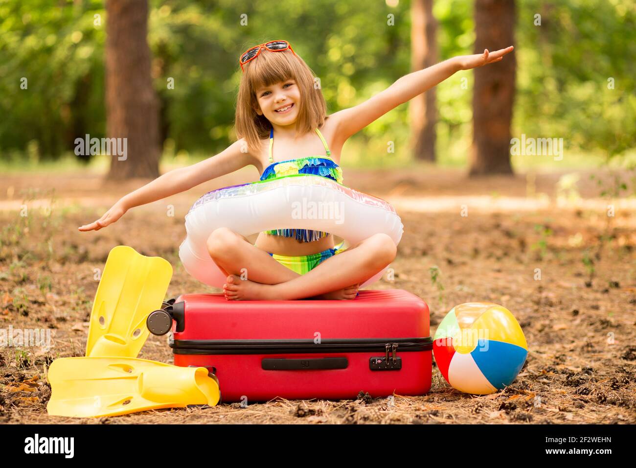 Little child girl wear swimsuit sitting on suitcase in summer forest with hands up, show plane. Dream journey. Vacation concept Stock Photo
