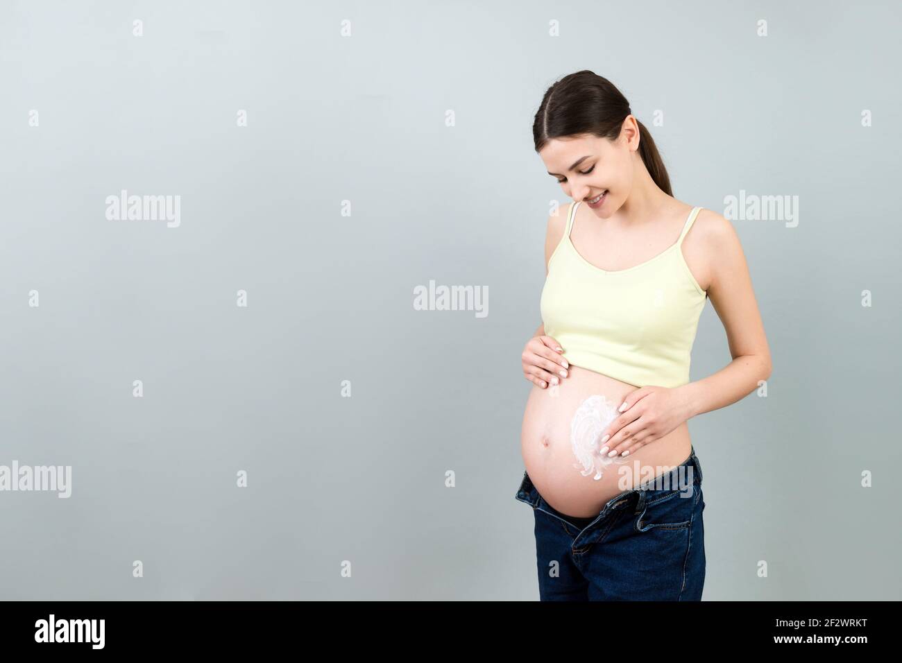 Pregnant Woman In Unzipped Jeans Applying Moisturizing Cream On Her Belly Against Stretch Marks 