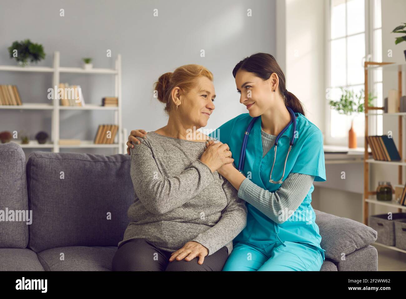 Happy doctor and senior patient sitting on couch, holding hands and looking at each other Stock
