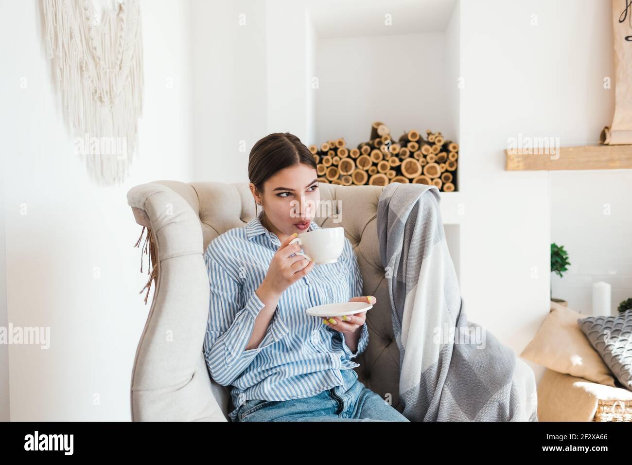 Young caucasian woman sitting on armchair in living room and drinking ...