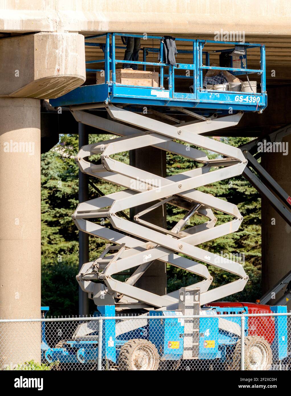 Scissor lift working under an overpass. A worker's legs can be seen on top of the lift. The bottom of the lift is behind a chain link fence. Stock Photo