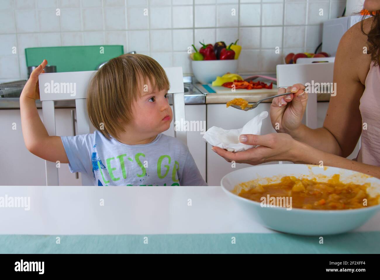 Mom feeding her disabled child with down syndrome. Mother giving food to toddler at home. Child doesn't like food. Stock Photo