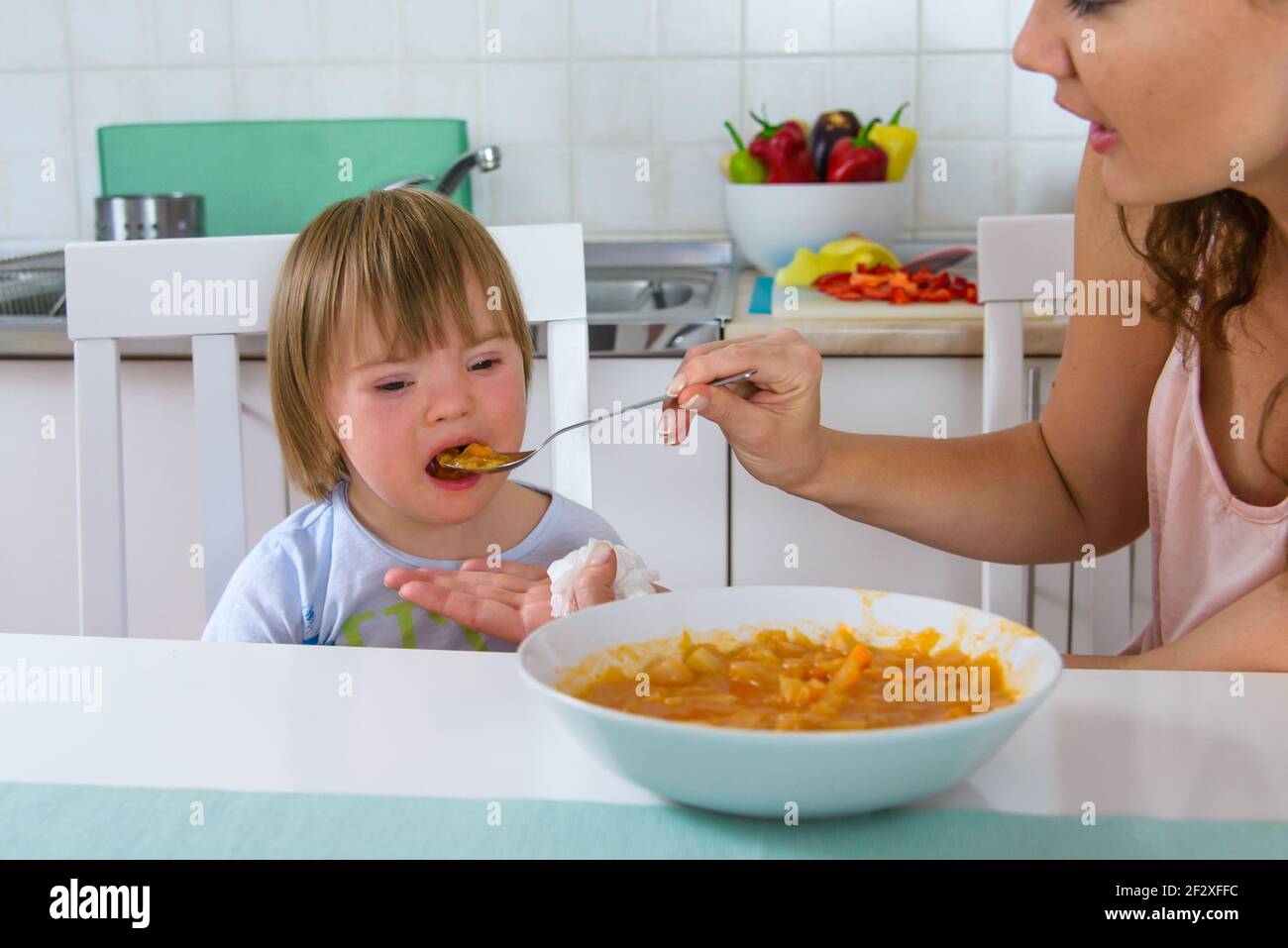 Mom feeding her disabled child with down syndrome with a spoon. Boy doesn't like offered food. Mother giving food to toddler at home. Baby food. Stock Photo