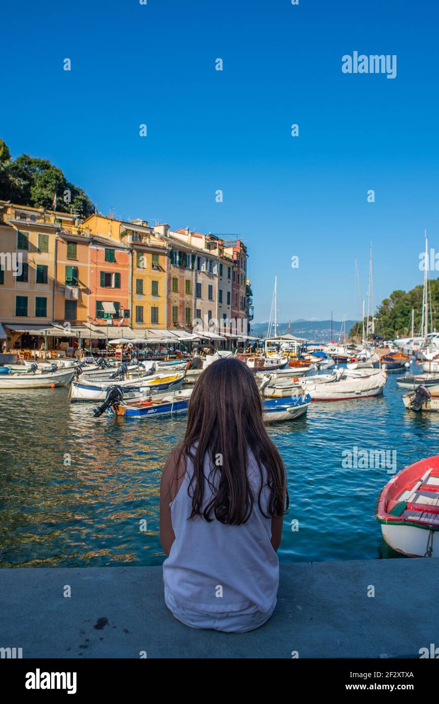 Young girl with long brown hair is sitting on a wall and looking at Portofino bay. The girl is caucasian and has a short sleeved shirt Stock Photo