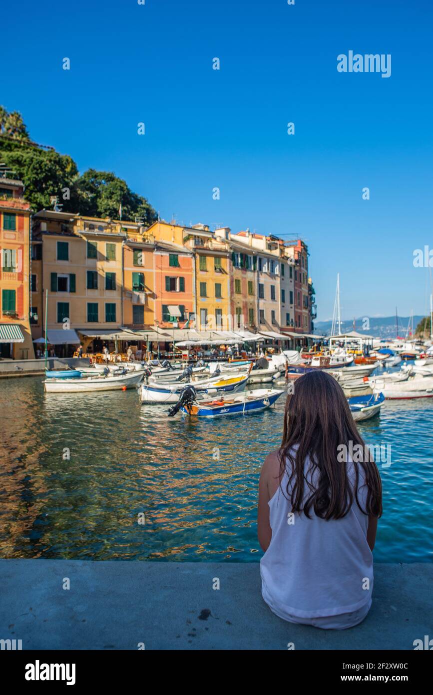 Young girl with long brown hair is sitting on a wall and looking at Portofino bay. The girl is caucasian and has a short sleeved shirt Stock Photo