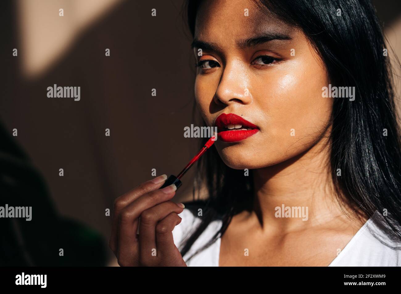 Portrait of crop thoughtful ethnic female with long dark hair looking at camera and rouging lips with red lipstick Stock Photo