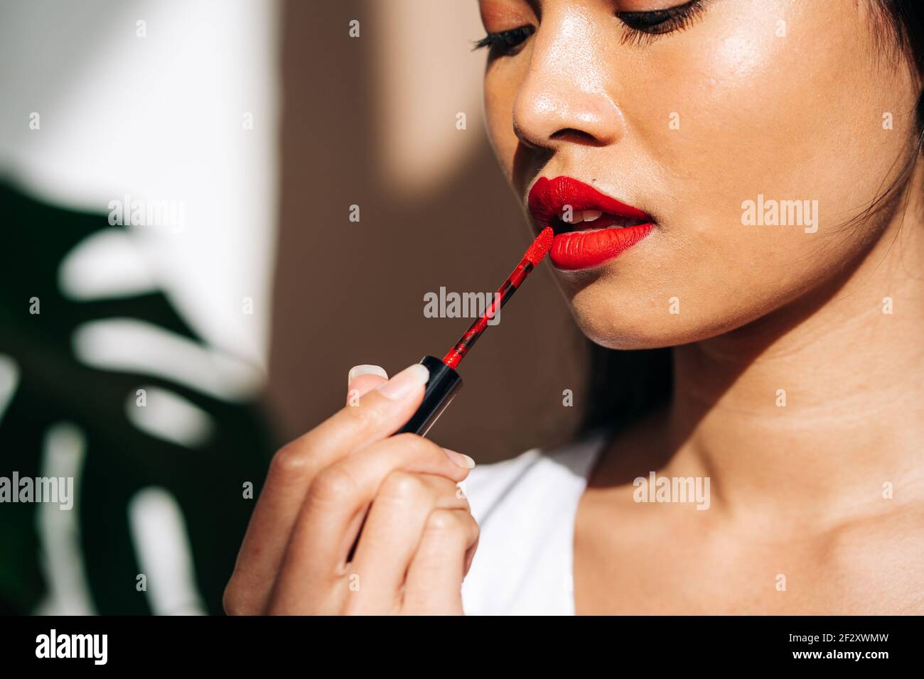 Portrait of crop thoughtful ethnic female with long dark hair looking down and rouging lips with red lipstick Stock Photo