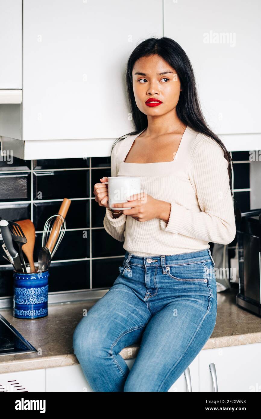 Wistful ethnic female in casual clothes sitting on countertop with cup of coffee while resting at home Stock Photo