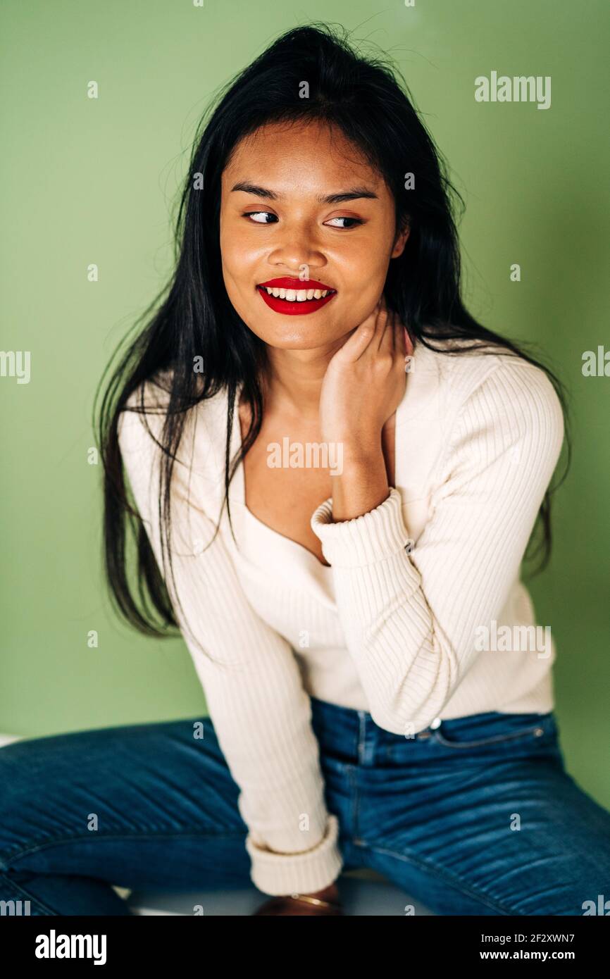 Attractive ethnic female model with bright lips looking at camera and leaning on hands sitting in studio on green background Stock Photo