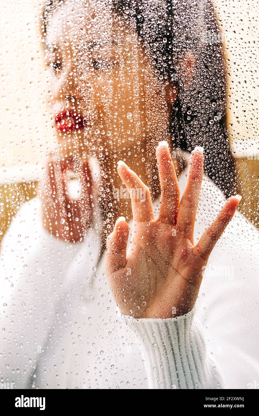 Through glass of pensive ethnic female with vivid lips looking away while touching drops Stock Photo