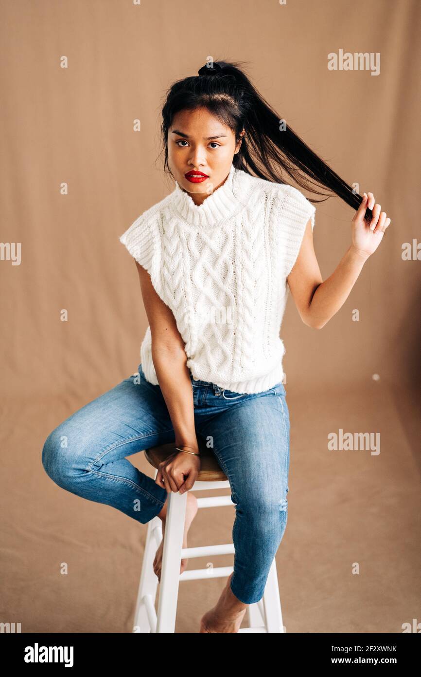 Confident young Hispanic lady wearing casual clothes sitting on wooden stool in studio Stock Photo