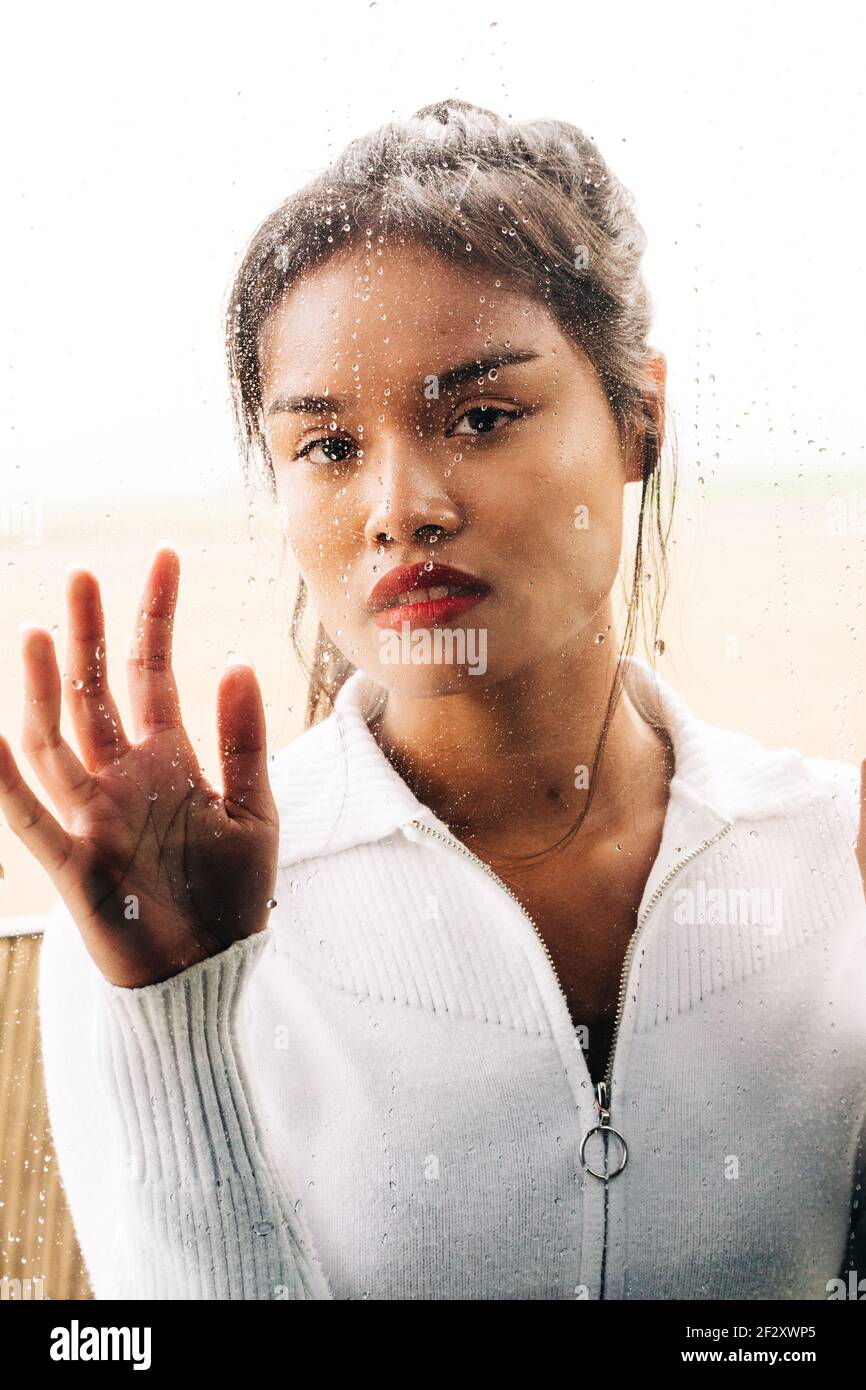 Through glass of pensive ethnic female with vivid lips looking at camera while touching drops Stock Photo