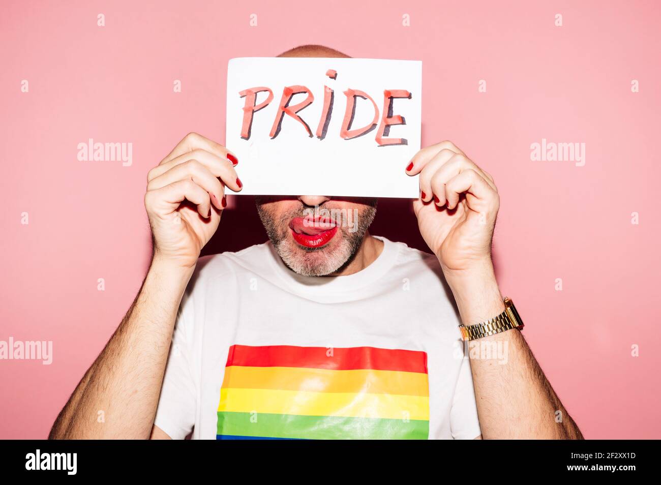 Rebellious bearded homosexual man with red lips and manicure making grimace with tongue out while showing and covering face with paper with Pride text Stock Photo