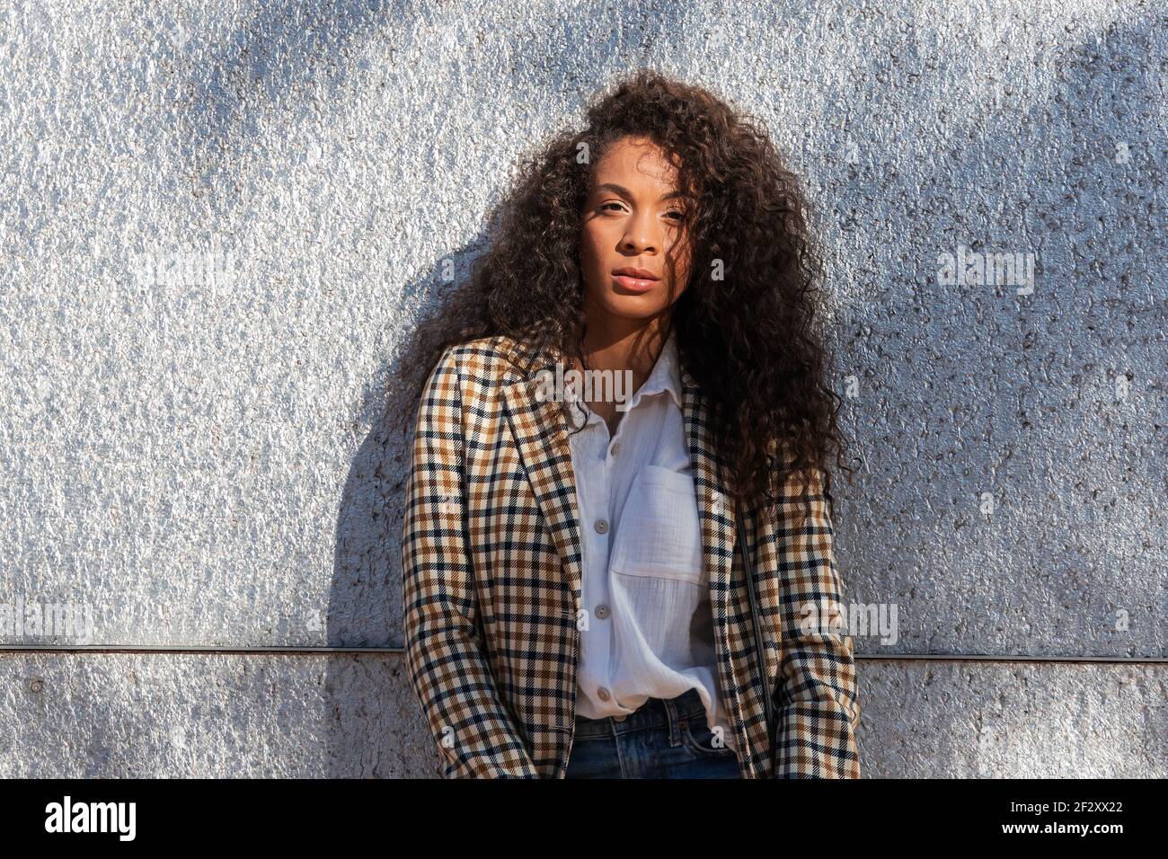 Pensive African American female in trendy outfit with curly hair looking at camera while standing on street near concrete wall Stock Photo