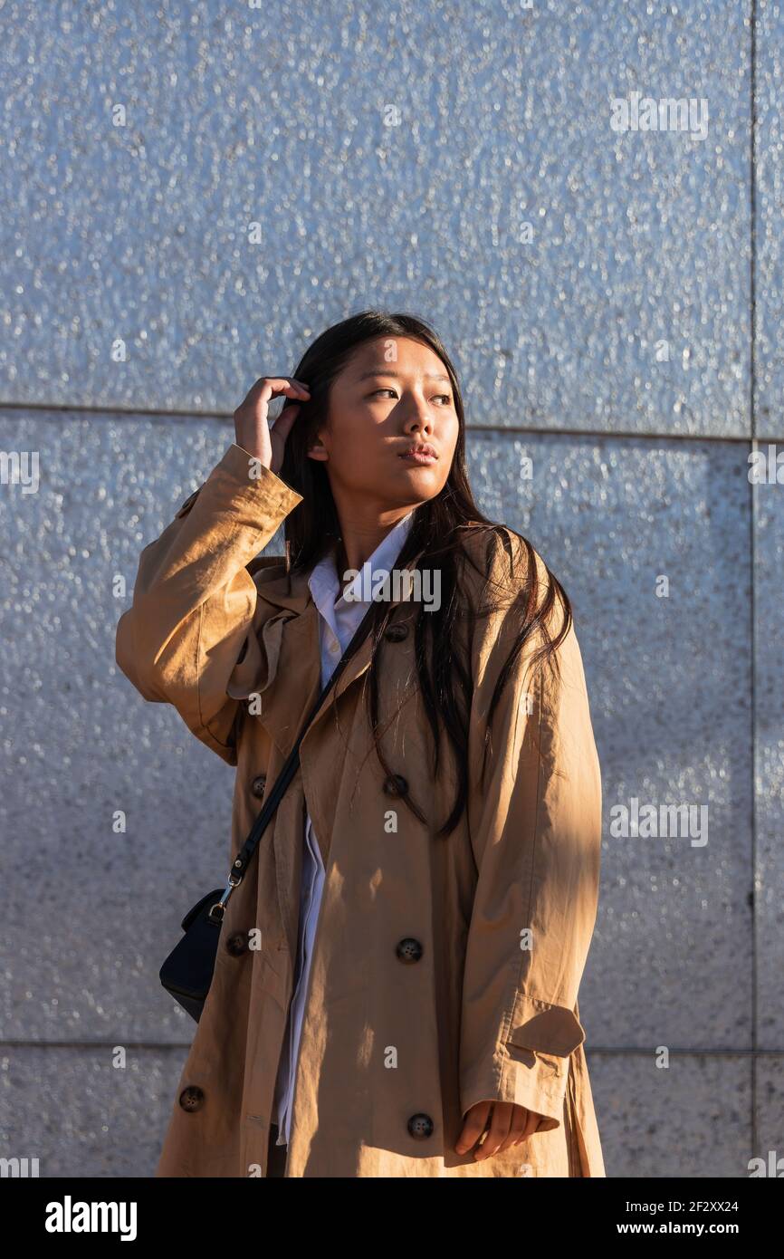 Serious Asian female in stylish outfit with long hair looking away while standing near modern building Stock Photo